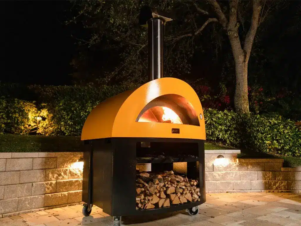 Forno a legna con cupola gialla su base in acciaio e legnaia, acceso in giardino